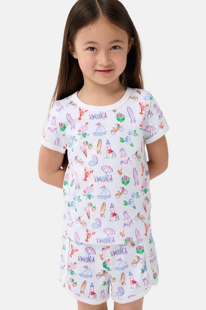 A young girl with long brown hair smiles, wearing the Roller Rabbit Kids Summer Club Playa Play Set made from soft Pima cotton, featuring colorful beach-themed illustrations on a plain light background.