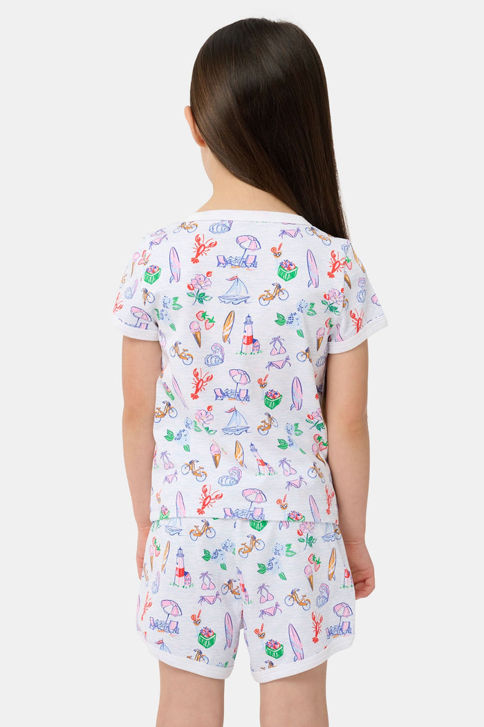 A young girl with long brown hair smiles, wearing the Roller Rabbit Kids Summer Club Playa Play Set made from soft Pima cotton, featuring colorful beach-themed illustrations on a plain light background.