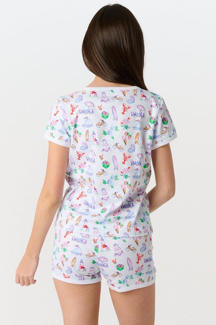A young girl with long brown hair smiles, wearing the Roller Rabbit Kids Summer Club Playa Play Set made from soft Pima cotton, featuring colorful beach-themed illustrations on a plain light background.