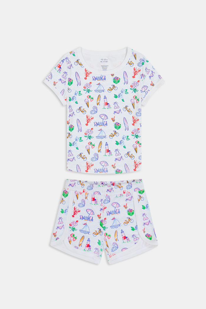 A young girl with long brown hair smiles, wearing the Roller Rabbit Kids Summer Club Playa Play Set made from soft Pima cotton, featuring colorful beach-themed illustrations on a plain light background.