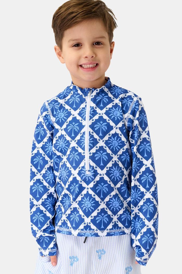 A young child with brown hair smiles, wearing the Roller Rabbit Kids Sunny Days Rashguard and light blue printed shorts, standing against a plain white background.