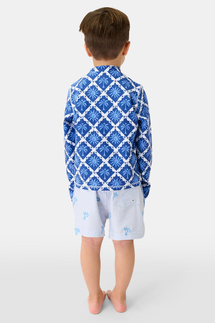 A young child with brown hair smiles, wearing the Roller Rabbit Kids Sunny Days Rashguard and light blue printed shorts, standing against a plain white background.