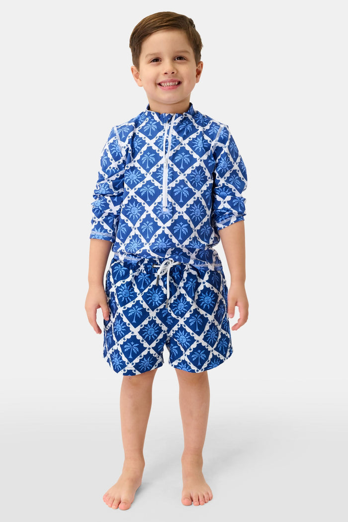The Roller Rabbit Boys Sunny Days Swim Trunk features a bold white lattice, blue floral and leaf motifs, white drawstring, adjustable waistband, and relaxed fit. Boys swimwear shorts are shown on a plain white background.