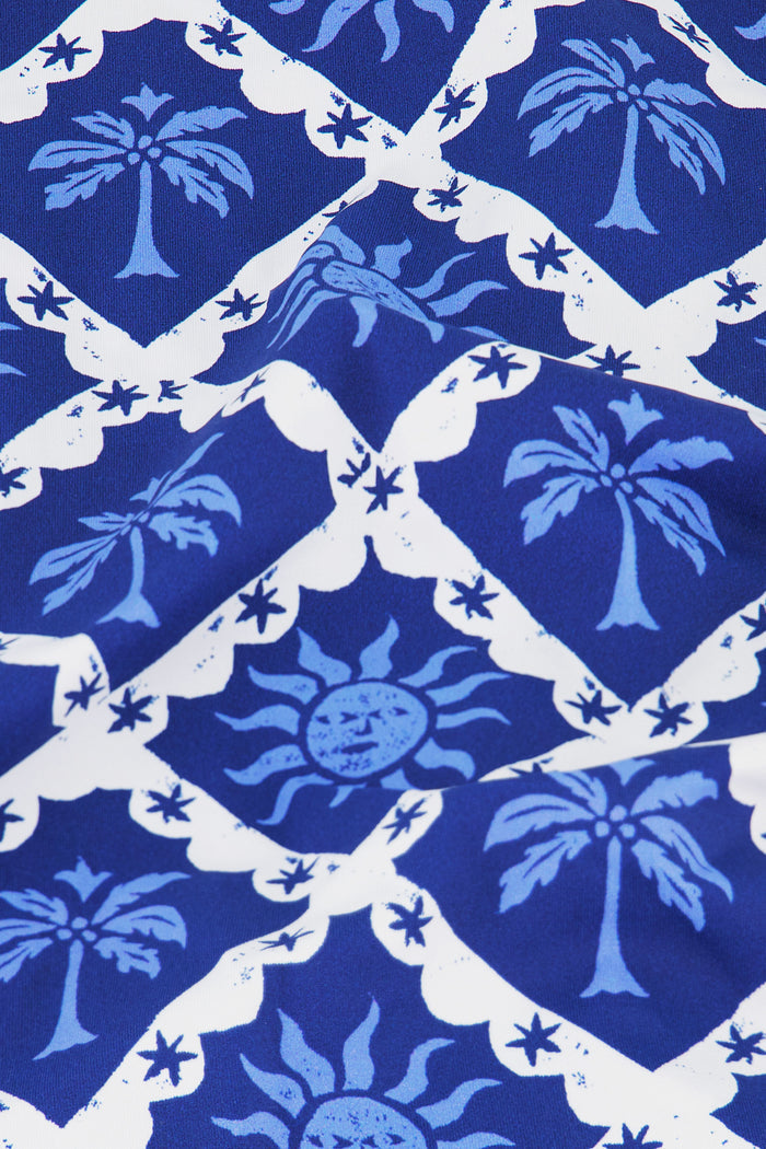 A young girl with long brown hair smiles, wearing the Kids Sunny Days Cora Bikini by Roller Rabbit—a blue and white patterned two-piece swimsuit with adjustable shoulder tie straps—standing against a plain light background.