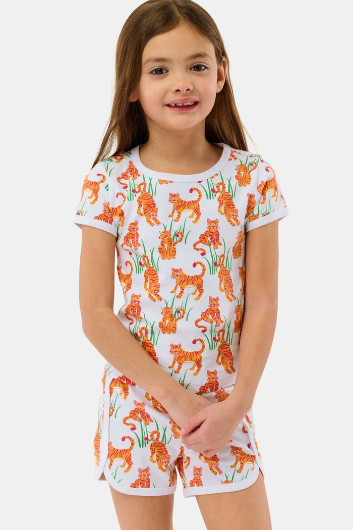 A young girl with long brown hair smiles in the Roller Rabbit Kids Teddy Tiger Playa Play Set, a white 100 Pima cotton outfit featuring colorful orange tiger and green plant patterns, standing against a plain light background.