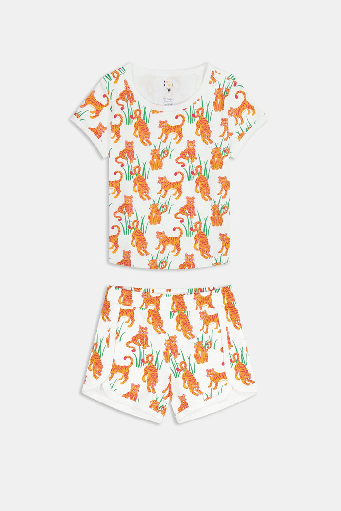 A young girl with long brown hair smiles in the Roller Rabbit Kids Teddy Tiger Playa Play Set, a white 100 Pima cotton outfit featuring colorful orange tiger and green plant patterns, standing against a plain light background.