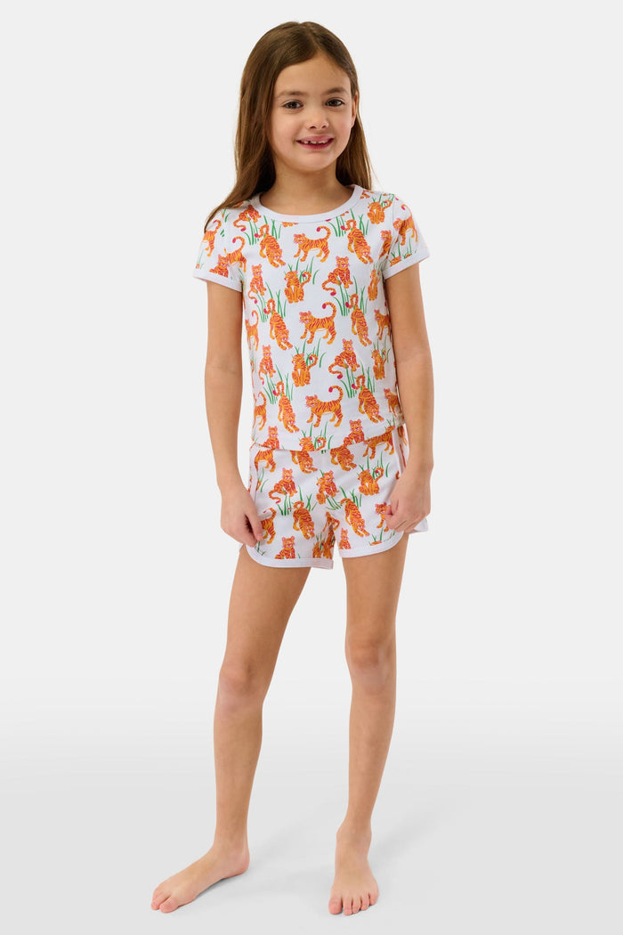 A young girl with long brown hair smiles in the Roller Rabbit Kids Teddy Tiger Playa Play Set, a white 100 Pima cotton outfit featuring colorful orange tiger and green plant patterns, standing against a plain light background.