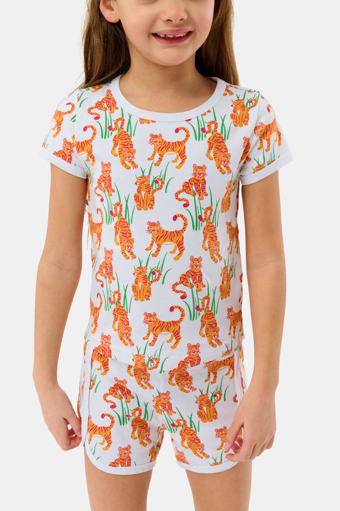 A young girl with long brown hair smiles in the Roller Rabbit Kids Teddy Tiger Playa Play Set, a white 100 Pima cotton outfit featuring colorful orange tiger and green plant patterns, standing against a plain light background.