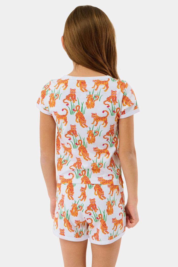 A young girl with long brown hair smiles in the Roller Rabbit Kids Teddy Tiger Playa Play Set, a white 100 Pima cotton outfit featuring colorful orange tiger and green plant patterns, standing against a plain light background.