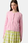 A woman with long brown hair models the Roller Rabbit Laine Cardigan—a cotton candy pink style with white buttons and front pockets—paired with a white and yellow plaid skirt with floral accents, set against a plain light background.