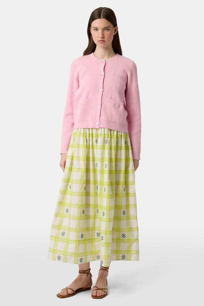 A woman with long brown hair models the Roller Rabbit Laine Cardigan—a cotton candy pink style with white buttons and front pockets—paired with a white and yellow plaid skirt with floral accents, set against a plain light background.