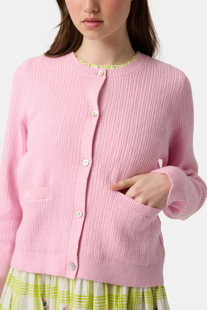 A woman with long brown hair models the Roller Rabbit Laine Cardigan—a cotton candy pink style with white buttons and front pockets—paired with a white and yellow plaid skirt with floral accents, set against a plain light background.