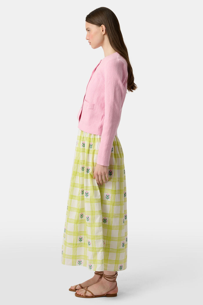 A woman with long brown hair models the Roller Rabbit Laine Cardigan—a cotton candy pink style with white buttons and front pockets—paired with a white and yellow plaid skirt with floral accents, set against a plain light background.