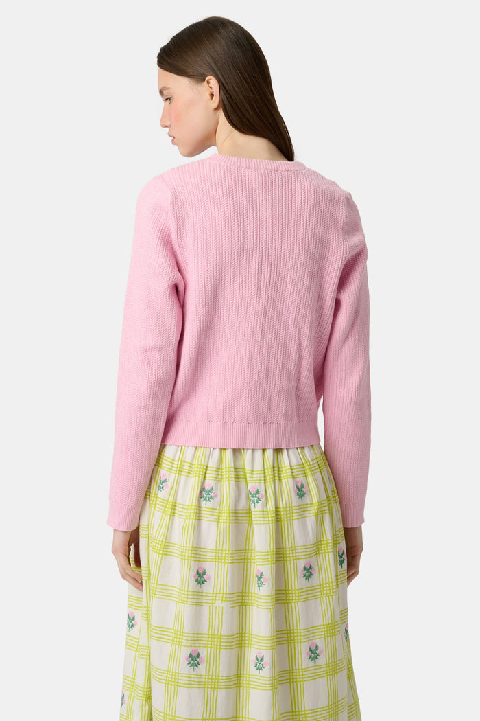 A woman with long brown hair models the Roller Rabbit Laine Cardigan—a cotton candy pink style with white buttons and front pockets—paired with a white and yellow plaid skirt with floral accents, set against a plain light background.