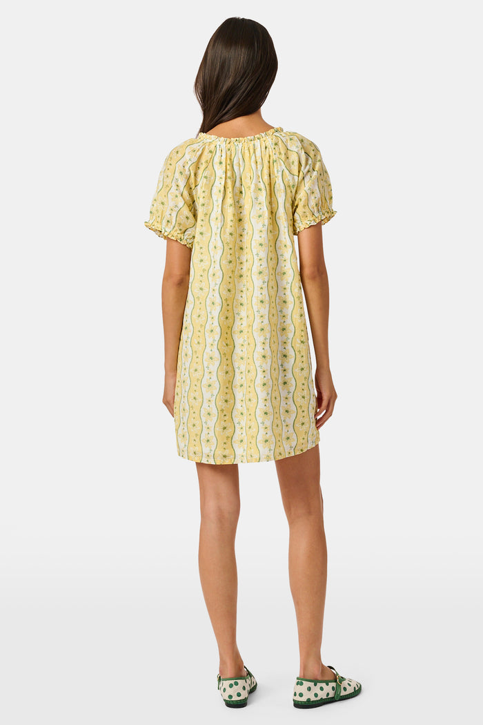 A girl wears the Roller Rabbit Kids Lillian Floral Stripe Tristan Dress, a cotton sundress with green and white stripes, floral patterns, ruffled straps, and a ruffled hem, standing with a smile and one arm relaxed at her side.