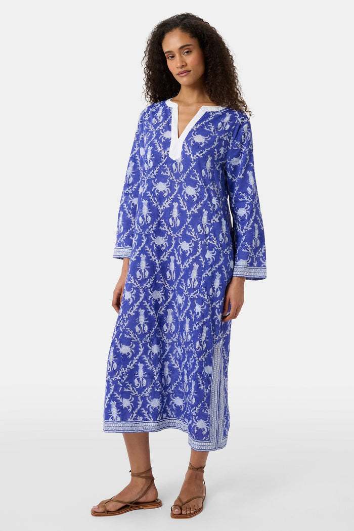 A woman wears the Roller Rabbit Lobster Trellis Johanna Caftan in blue and white, featuring a white V-neckline and hem, paired with sandals against a plain background.
