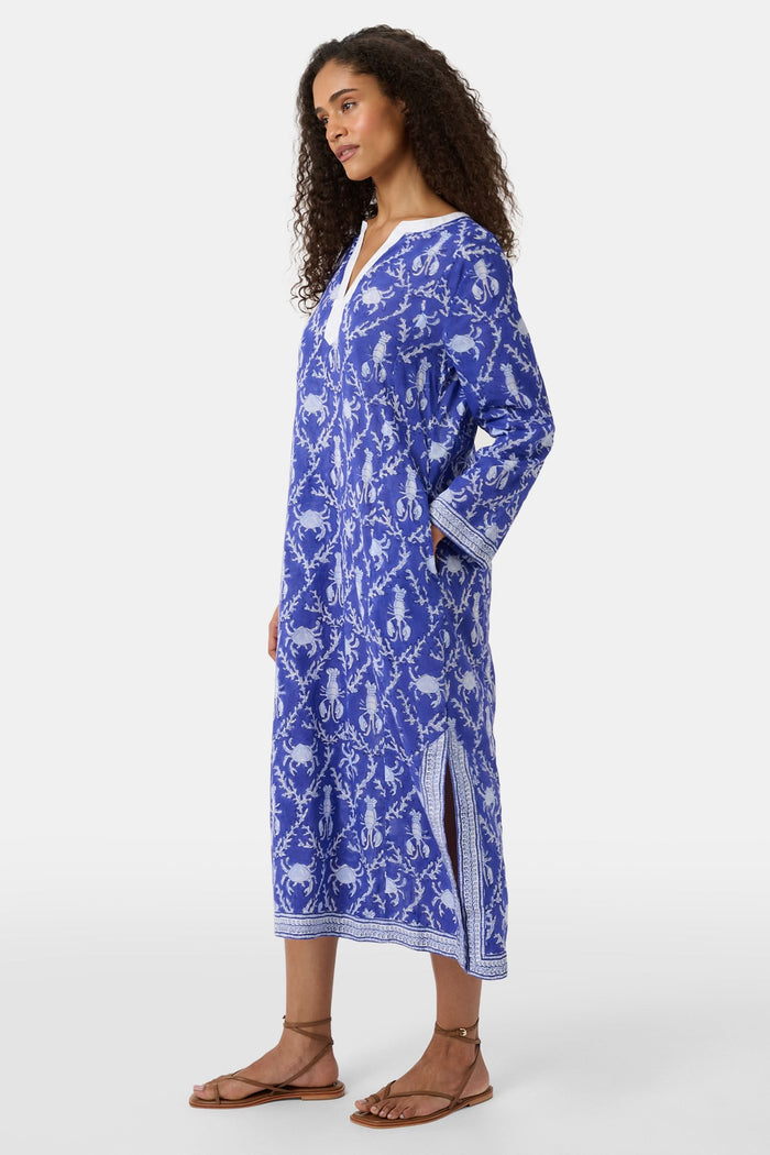 A woman wears the Roller Rabbit Lobster Trellis Johanna Caftan in blue and white, featuring a white V-neckline and hem, paired with sandals against a plain background.