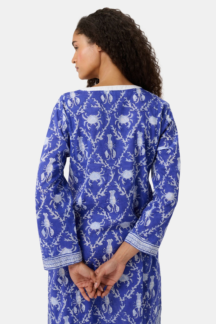A woman wears the Roller Rabbit Lobster Trellis Johanna Caftan in blue and white, featuring a white V-neckline and hem, paired with sandals against a plain background.