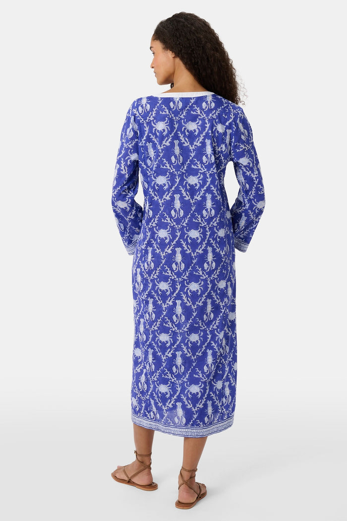 A woman wears the Roller Rabbit Lobster Trellis Johanna Caftan in blue and white, featuring a white V-neckline and hem, paired with sandals against a plain background.