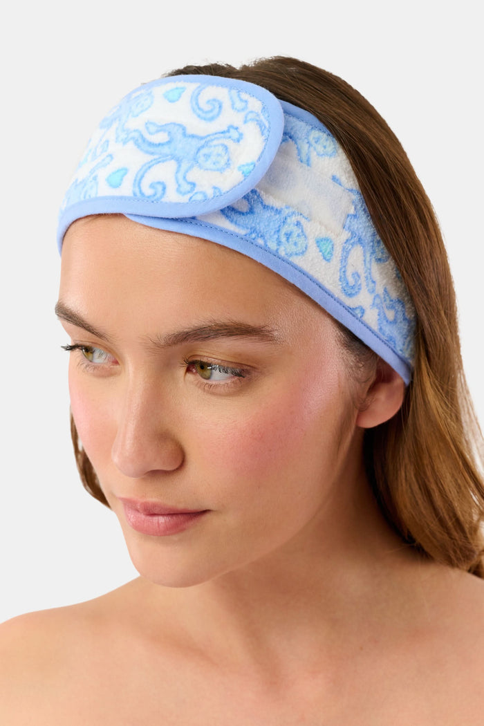 A woman with long brown hair wears the Roller Rabbit Lovely Monkey Spa Headband, a blue and white patterned terry accessory, pushed up on her forehead. She has clear skin, a slight smile, and stands against a plain white background.
