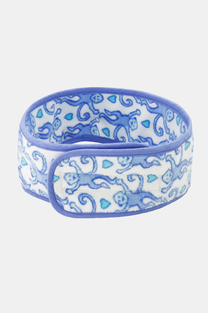 A woman with long brown hair wears the Roller Rabbit Lovely Monkey Spa Headband, a blue and white patterned terry accessory, pushed up on her forehead. She has clear skin, a slight smile, and stands against a plain white background.