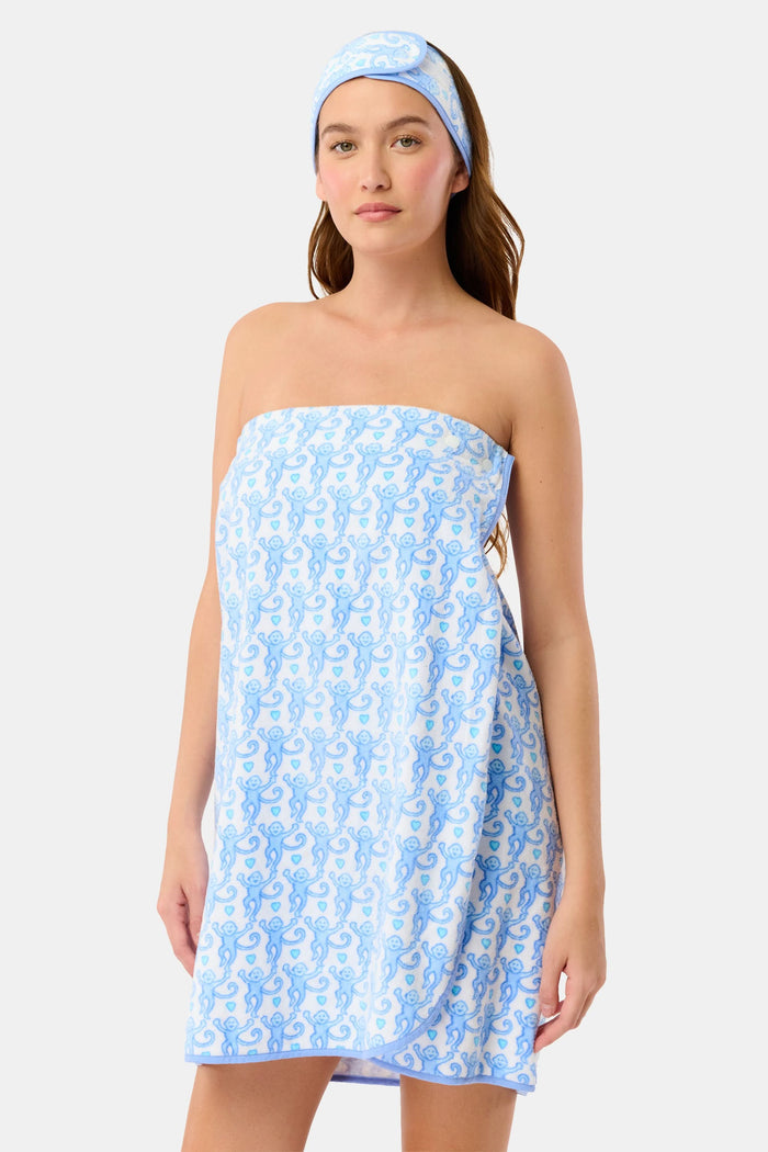 A woman with long dark hair stands in a doorway wearing the Roller Rabbit Lovely Monkey Towel Wrap and matching headband, gazing to the side with her hand on her chest and a relaxed, thoughtful expression.
