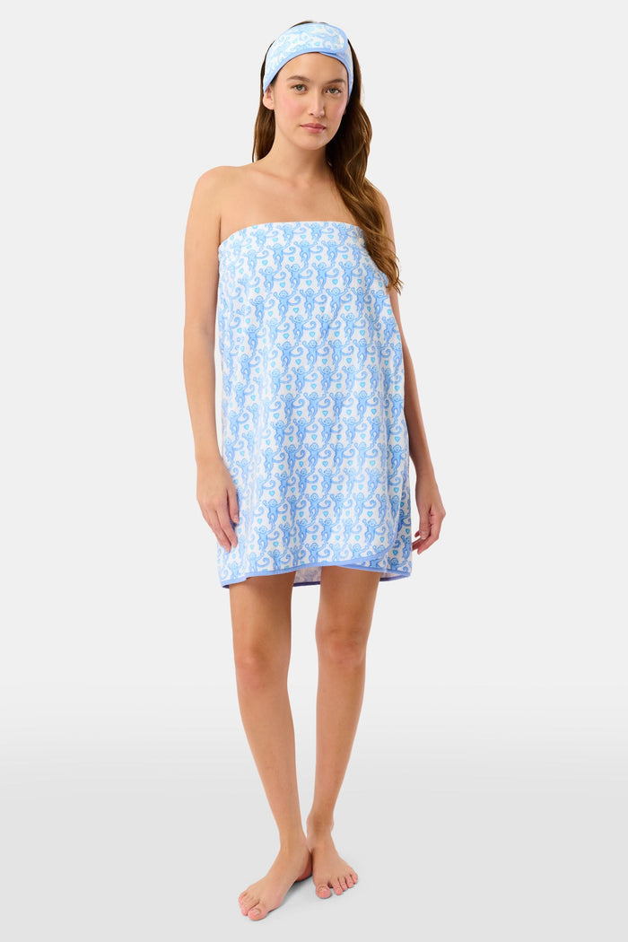 A woman with long dark hair stands in a doorway wearing the Roller Rabbit Lovely Monkey Towel Wrap and matching headband, gazing to the side with her hand on her chest and a relaxed, thoughtful expression.