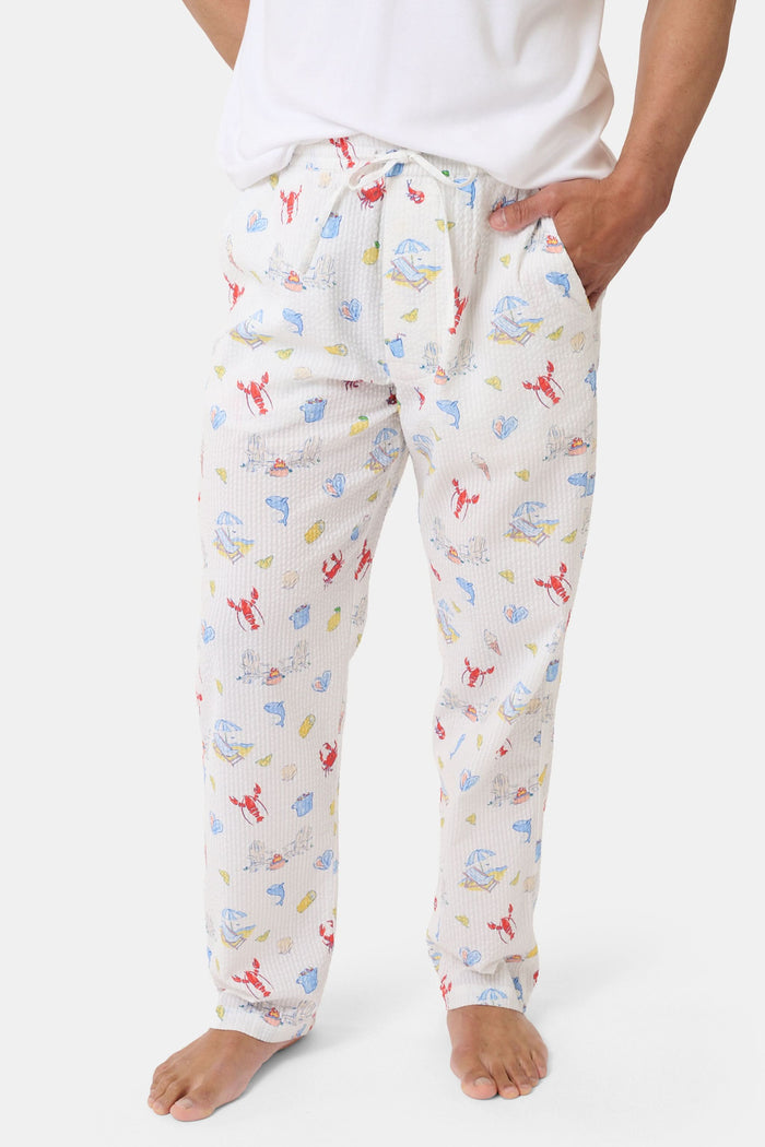 A person stands barefoot in Roller Rabbit’s Men’s Catch of the Day Jon Pant—loose white seersucker pants with a lobsters, fish, and seashells print—and a white t-shirt, one hand tucked into the pants’ pocket.