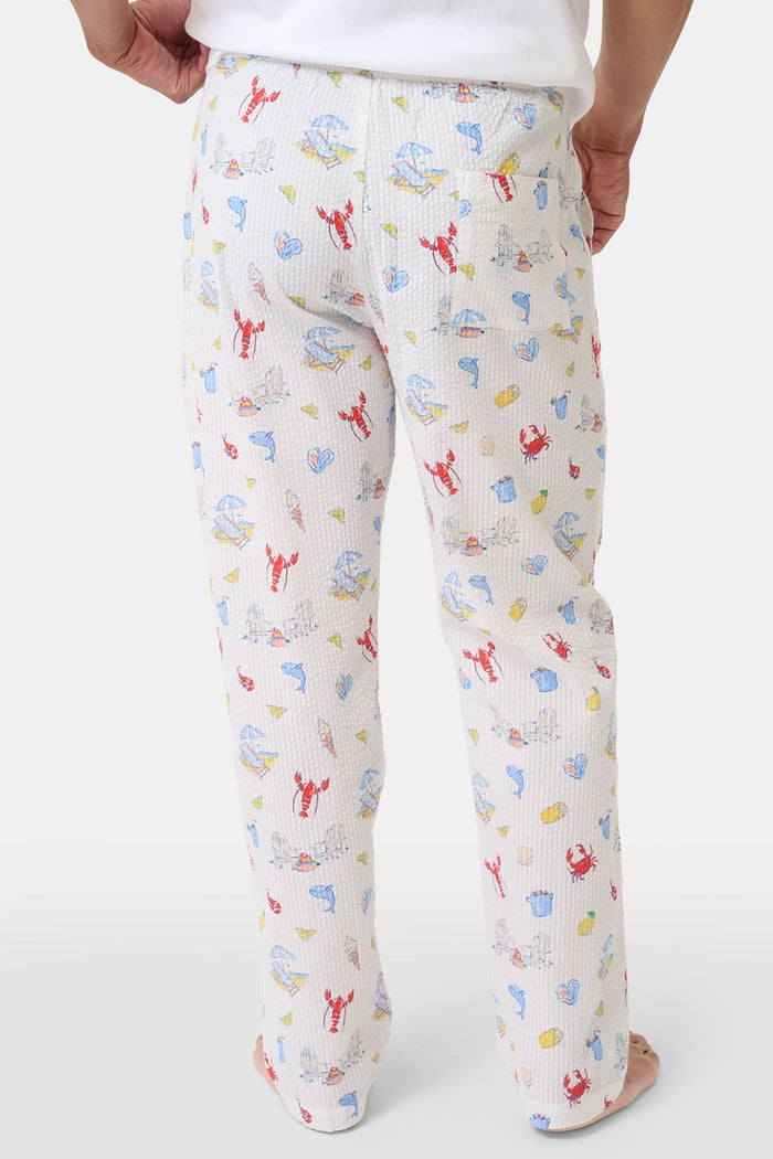 A person stands barefoot in Roller Rabbit’s Men’s Catch of the Day Jon Pant—loose white seersucker pants with a lobsters, fish, and seashells print—and a white t-shirt, one hand tucked into the pants’ pocket.