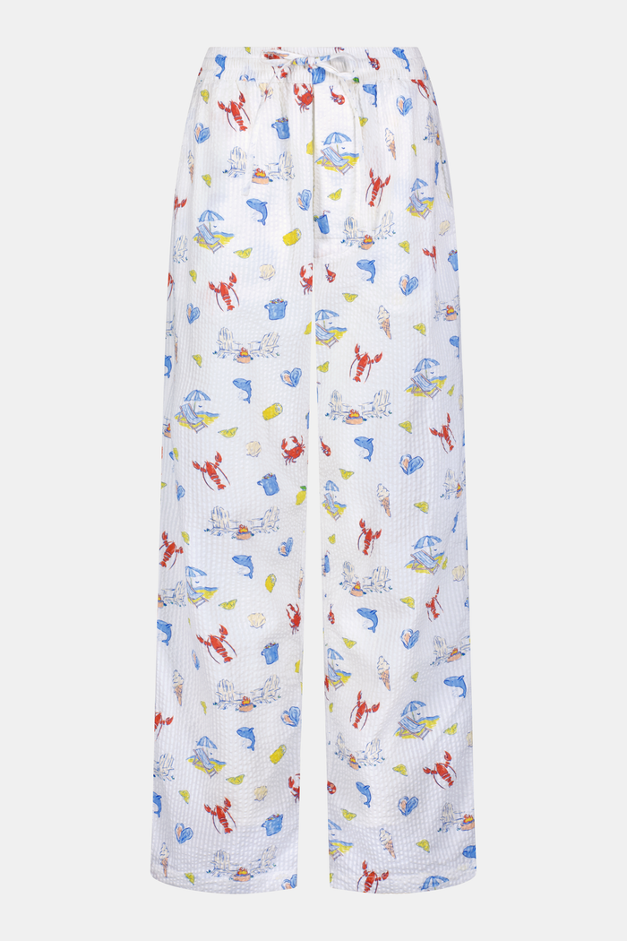 A person stands barefoot in Roller Rabbit’s Men’s Catch of the Day Jon Pant—loose white seersucker pants with a lobsters, fish, and seashells print—and a white t-shirt, one hand tucked into the pants’ pocket.