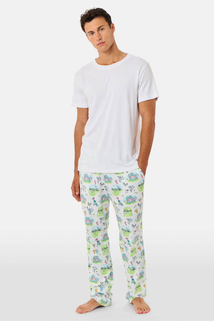 A person wearing Roller Rabbit Mens Game, Set, Monkey Troy Pajamas—white Pima Cotton pajama pants featuring a playful camp-themed print with tents, trees, and campfires—paired with a white T-shirt, standing barefoot against a plain background.