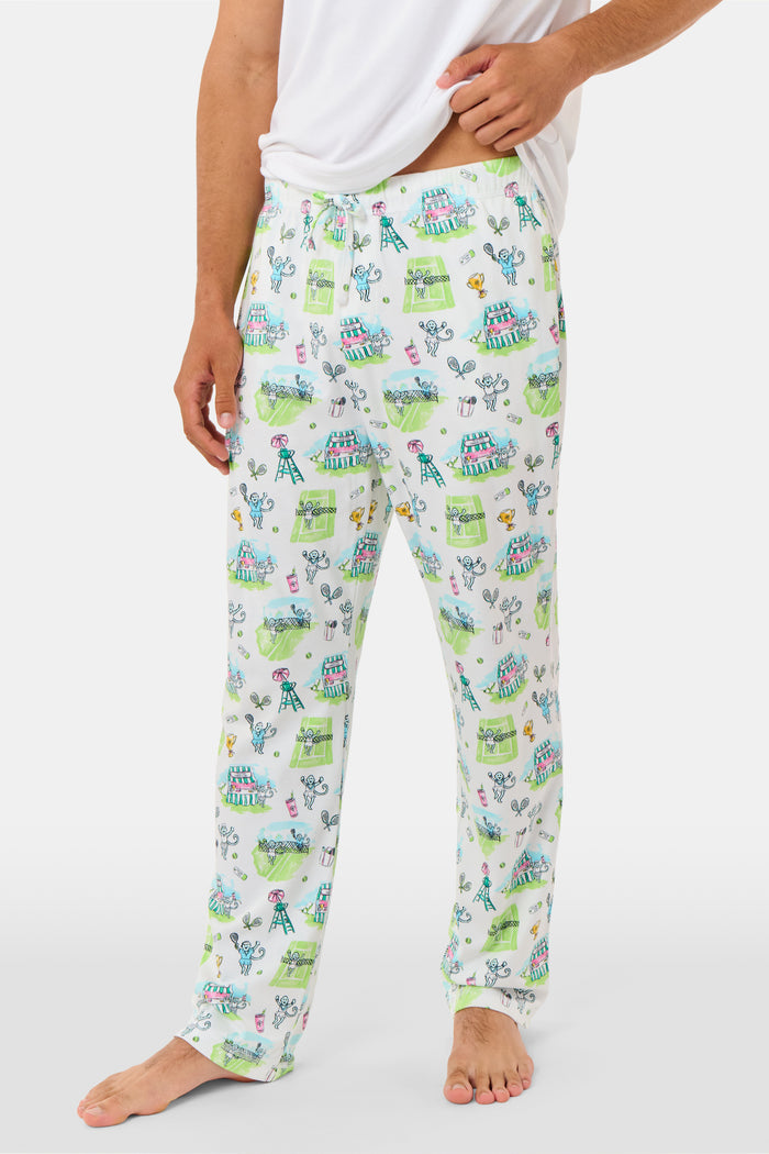 A person wearing Roller Rabbit Mens Game, Set, Monkey Troy Pajamas—white Pima Cotton pajama pants featuring a playful camp-themed print with tents, trees, and campfires—paired with a white T-shirt, standing barefoot against a plain background.