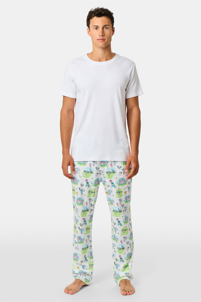 A person wearing Roller Rabbit Mens Game, Set, Monkey Troy Pajamas—white Pima Cotton pajama pants featuring a playful camp-themed print with tents, trees, and campfires—paired with a white T-shirt, standing barefoot against a plain background.