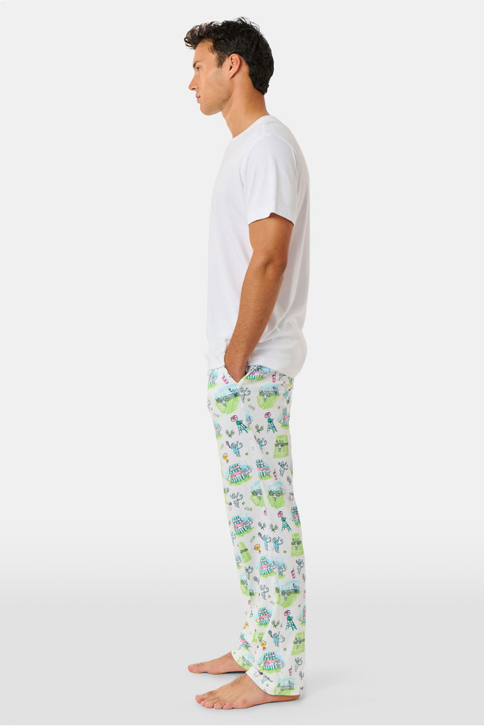 A person wearing Roller Rabbit Mens Game, Set, Monkey Troy Pajamas—white Pima Cotton pajama pants featuring a playful camp-themed print with tents, trees, and campfires—paired with a white T-shirt, standing barefoot against a plain background.
