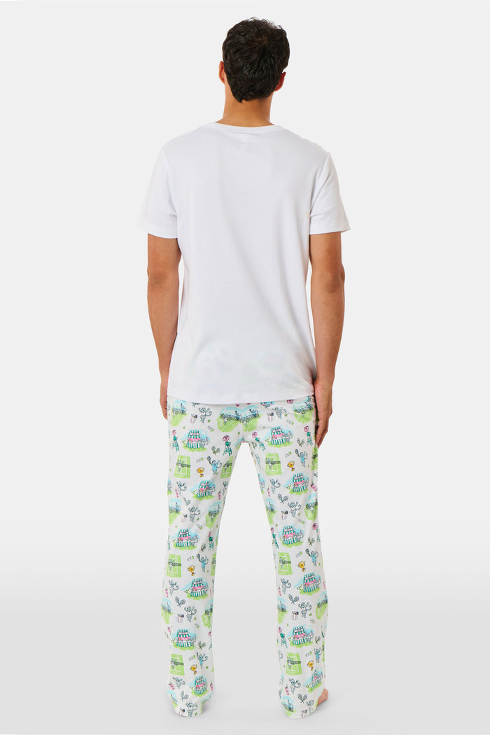 A person wearing Roller Rabbit Mens Game, Set, Monkey Troy Pajamas—white Pima Cotton pajama pants featuring a playful camp-themed print with tents, trees, and campfires—paired with a white T-shirt, standing barefoot against a plain background.