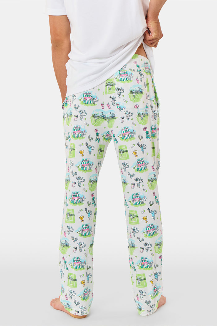 A person wearing Roller Rabbit Mens Game, Set, Monkey Troy Pajamas—white Pima Cotton pajama pants featuring a playful camp-themed print with tents, trees, and campfires—paired with a white T-shirt, standing barefoot against a plain background.