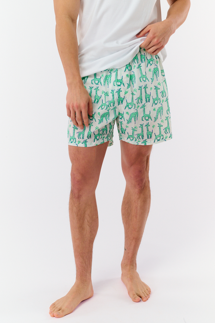 A man stands barefoot in a white T-shirt and Roller Rabbit’s Mens Gigi Giraffe Boxer Set, featuring green giraffe print shorts—ideal for showcasing these stylish cotton boxers as a thoughtful men’s gift.