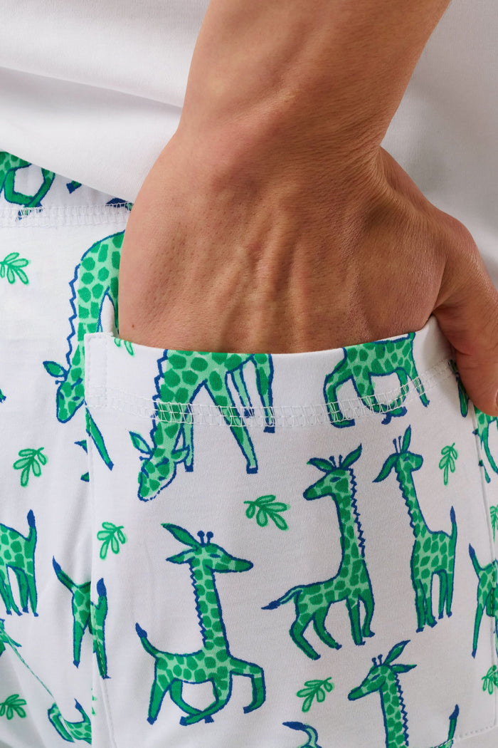 A person wears Roller Rabbit's Mens Gigi Giraffe Simon Pant, white pajama pants with a giraffe print, standing barefoot against a plain white background. Made from 100 Pima Cotton, the pull-on style features a hand in the pocket and a partially visible upper body.