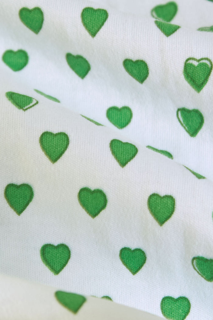 A woman smiles in Roller Rabbit’s Hearts Polo Pajamas—a white, short-sleeve Pima cotton set with emerald green hearts, a collared button-up top, and matching shorts—standing against a plain white background.