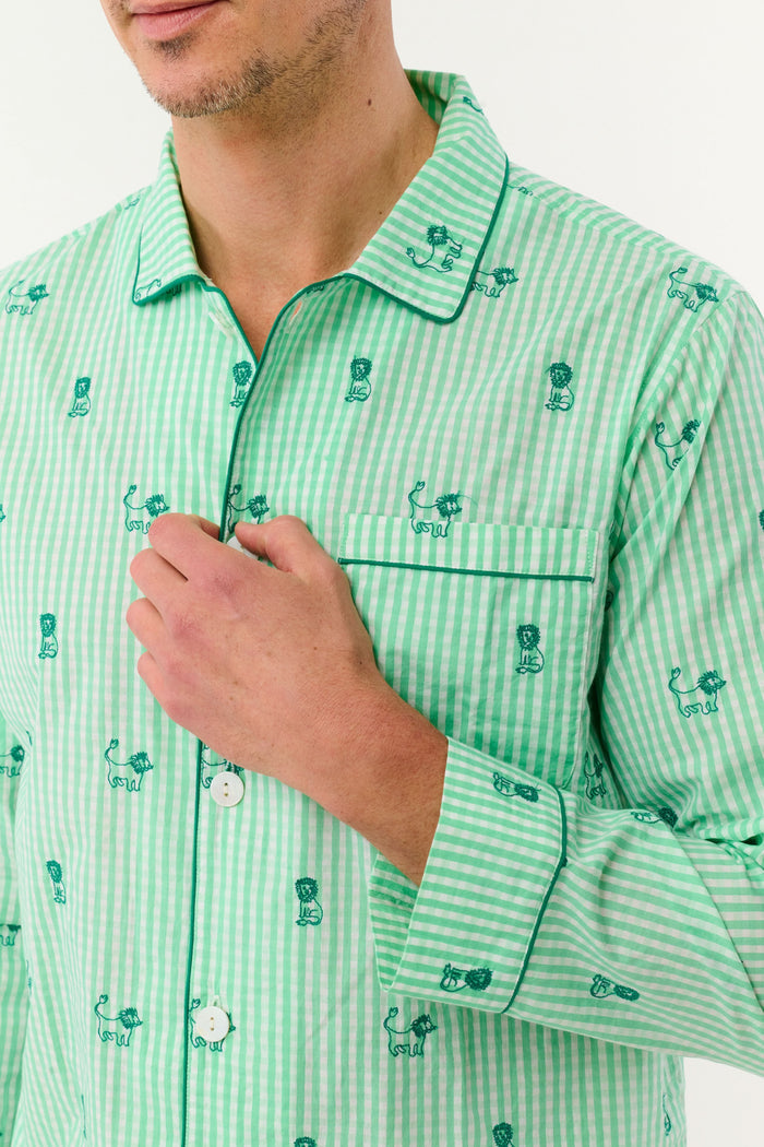A man stands barefoot against a white background, smiling and facing forward, wearing Roller Rabbit's Mens Leo Gingham Loungewear—a light green, long-sleeved button-up set with matching pants featuring a small lion print pattern.
