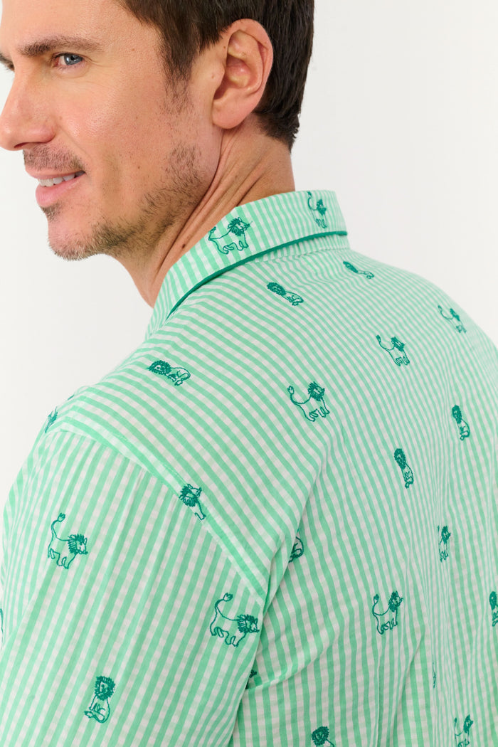 A man stands barefoot against a white background, smiling and facing forward, wearing Roller Rabbit's Mens Leo Gingham Loungewear—a light green, long-sleeved button-up set with matching pants featuring a small lion print pattern.