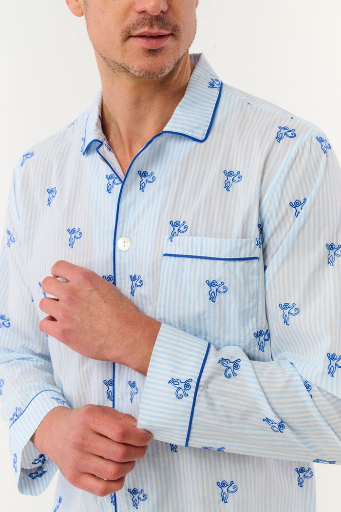 A man stands barefoot wearing the Roller Rabbit Mens Mini Monkey Stripe Loungewear—a cotton pajama set featuring light blue stripes with a small blue floral pattern, long sleeves, and matching pants. He looks at the camera with a slight smile against a plain white background, embodying cozy sleepwear comfort.
