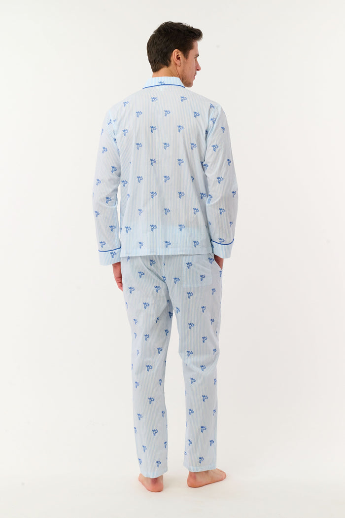 A man stands barefoot wearing the Roller Rabbit Mens Mini Monkey Stripe Loungewear—a cotton pajama set featuring light blue stripes with a small blue floral pattern, long sleeves, and matching pants. He looks at the camera with a slight smile against a plain white background, embodying cozy sleepwear comfort.