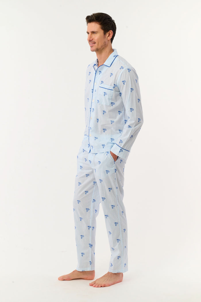 A man stands barefoot wearing the Roller Rabbit Mens Mini Monkey Stripe Loungewear—a cotton pajama set featuring light blue stripes with a small blue floral pattern, long sleeves, and matching pants. He looks at the camera with a slight smile against a plain white background, embodying cozy sleepwear comfort.