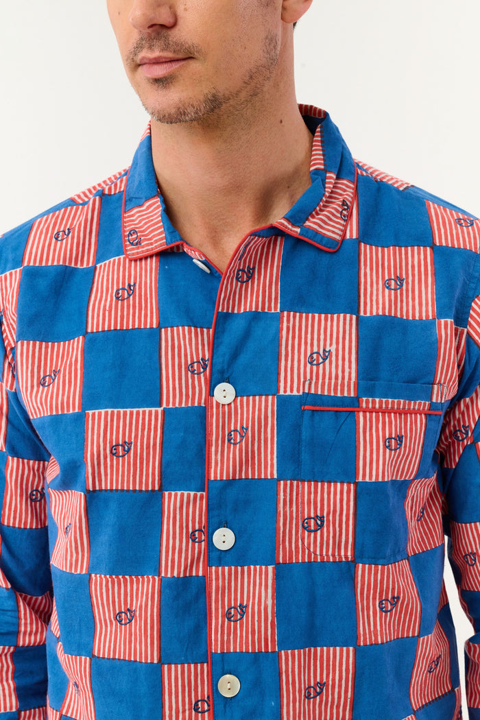 A man with short brown hair and a neutral expression stands barefoot against a white background, wearing the Roller Rabbit Mens Moby Grand Prix Loungewear set in blue and pink checkered cotton with long sleeves and pants.