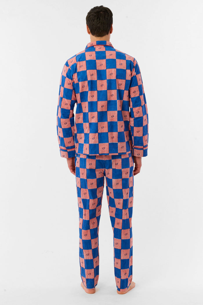 A man with short brown hair and a neutral expression stands barefoot against a white background, wearing the Roller Rabbit Mens Moby Grand Prix Loungewear set in blue and pink checkered cotton with long sleeves and pants.