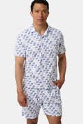 A man stands barefoot facing forward in Roller Rabbit’s Mens Moby Louie Pajamas—a Pima cotton set with blue patterns on white, featuring a short-sleeve button-up shirt and shorts—against a plain white background.