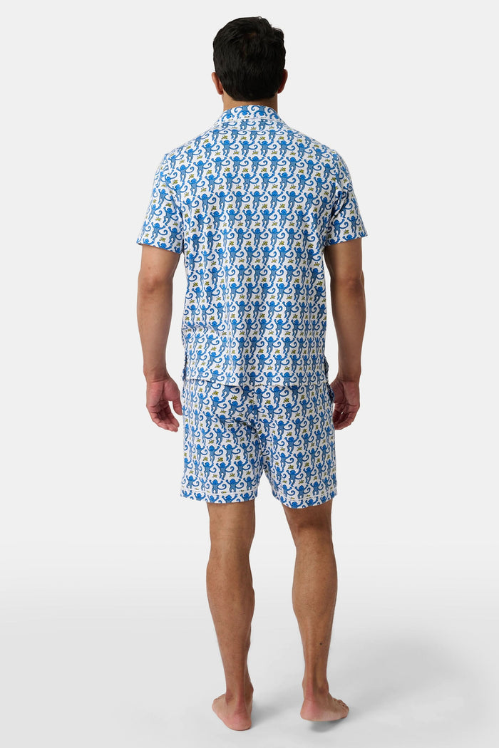 A man stands barefoot against a white background wearing Roller Rabbit Mens Monkey Louie Pajamas—a matching short-sleeve button-up shirt and shorts featuring a blue and white abstract print in 100 Pima cotton.