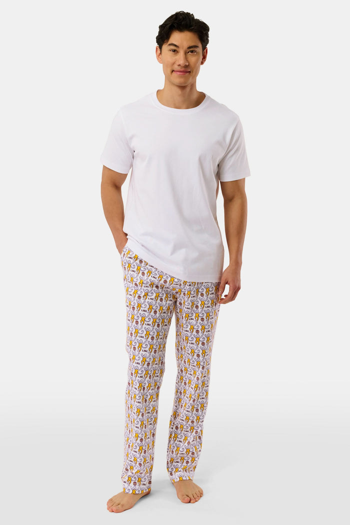 A man with short dark hair stands barefoot against a plain background, wearing a white t-shirt and Roller Rabbit Mens Tigers Lovely Monkey Simon Pant lounge pants with a yellow and brown design. He has a relaxed, neutral expression.