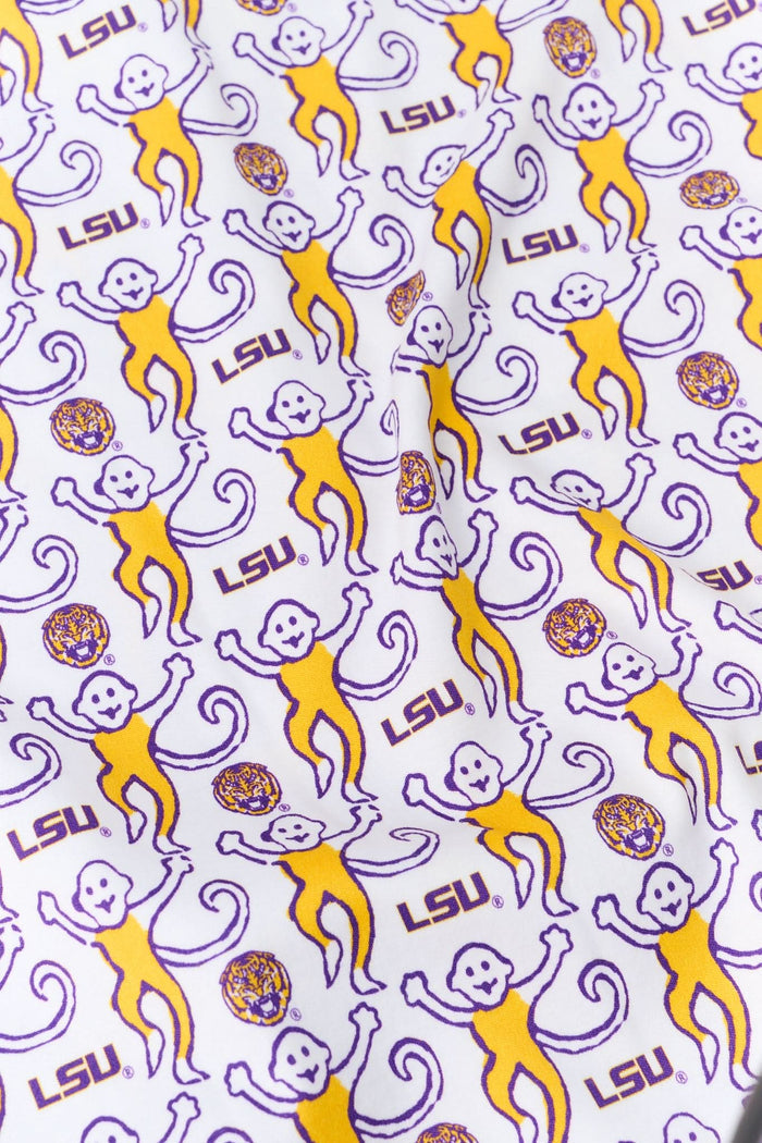 A woman stands against a plain background wearing Roller Rabbit’s Tigers Lovely Monkey Polo Pajamas, an LSU-themed set featuring a playful purple, yellow, and white abstract pattern with a short-sleeve button-up top and shorts.